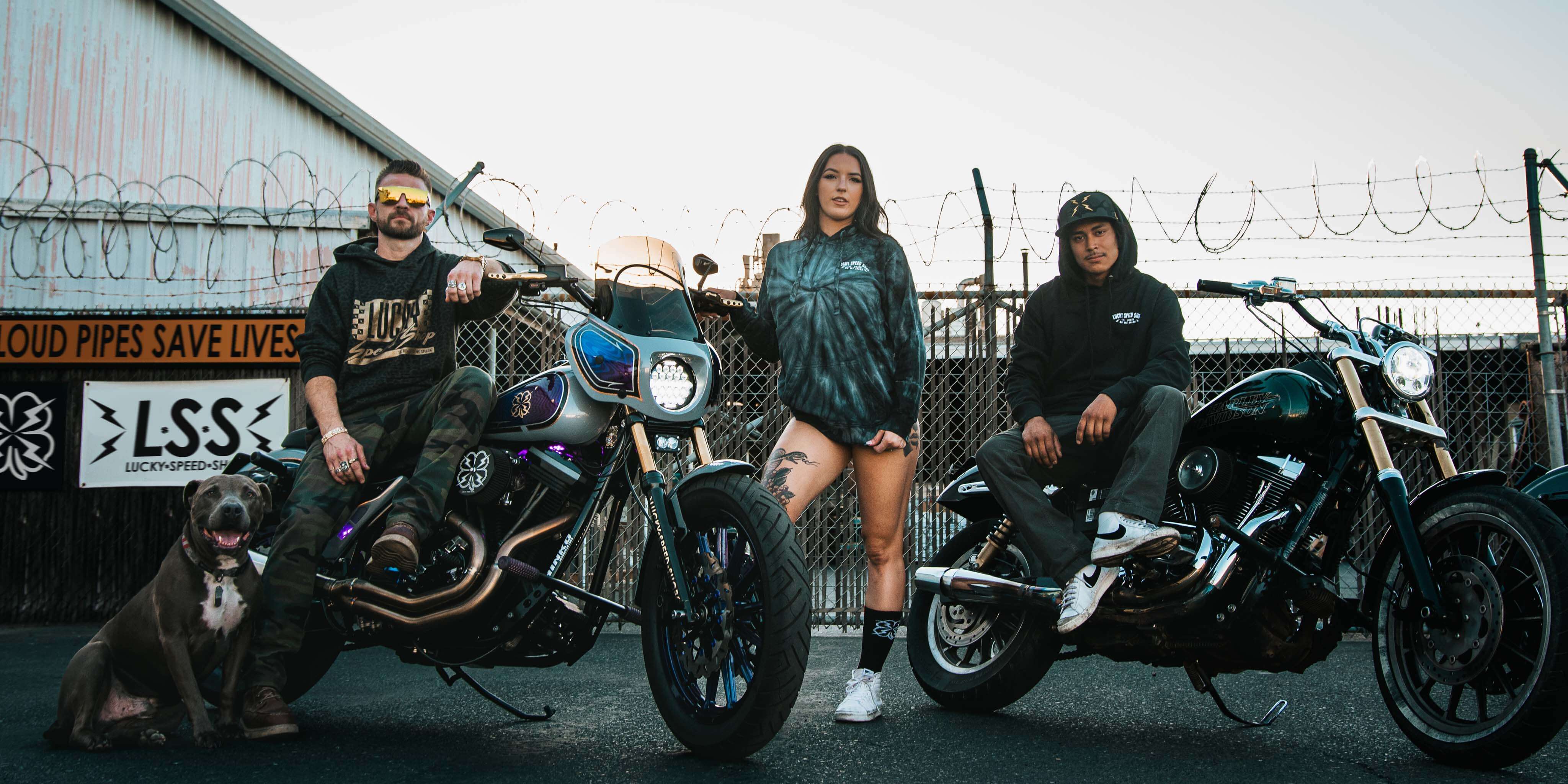 Lucky Speed Crew (including bluenose pitbull) sitting on 1986 Harley FXR and 2014 Harley Dyna street bob with woman model in the middle wearing lucky speed shop clothing